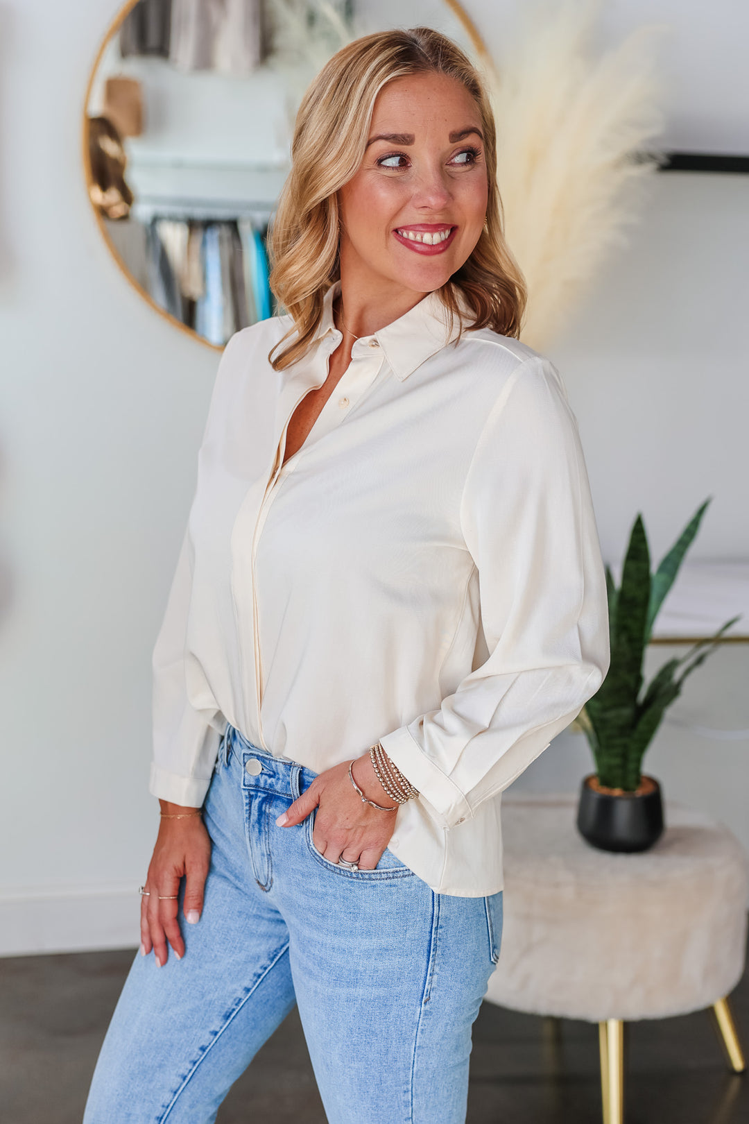 A blonde woman standing in a shop wearing a cream colored button down with blue jeans.