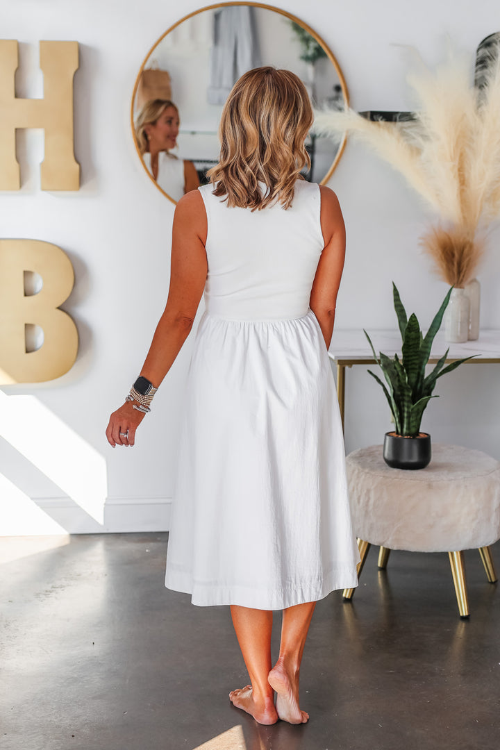 A blonde woman standing in a shop wearing a white dress with a zip collar closure and nylon skirt. She is rear facing.