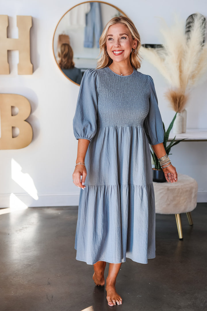 A blonde woman standing in a shop wearing a denim colored dress with 3/4 length sleeves, smocked bodice and tiered skirt.