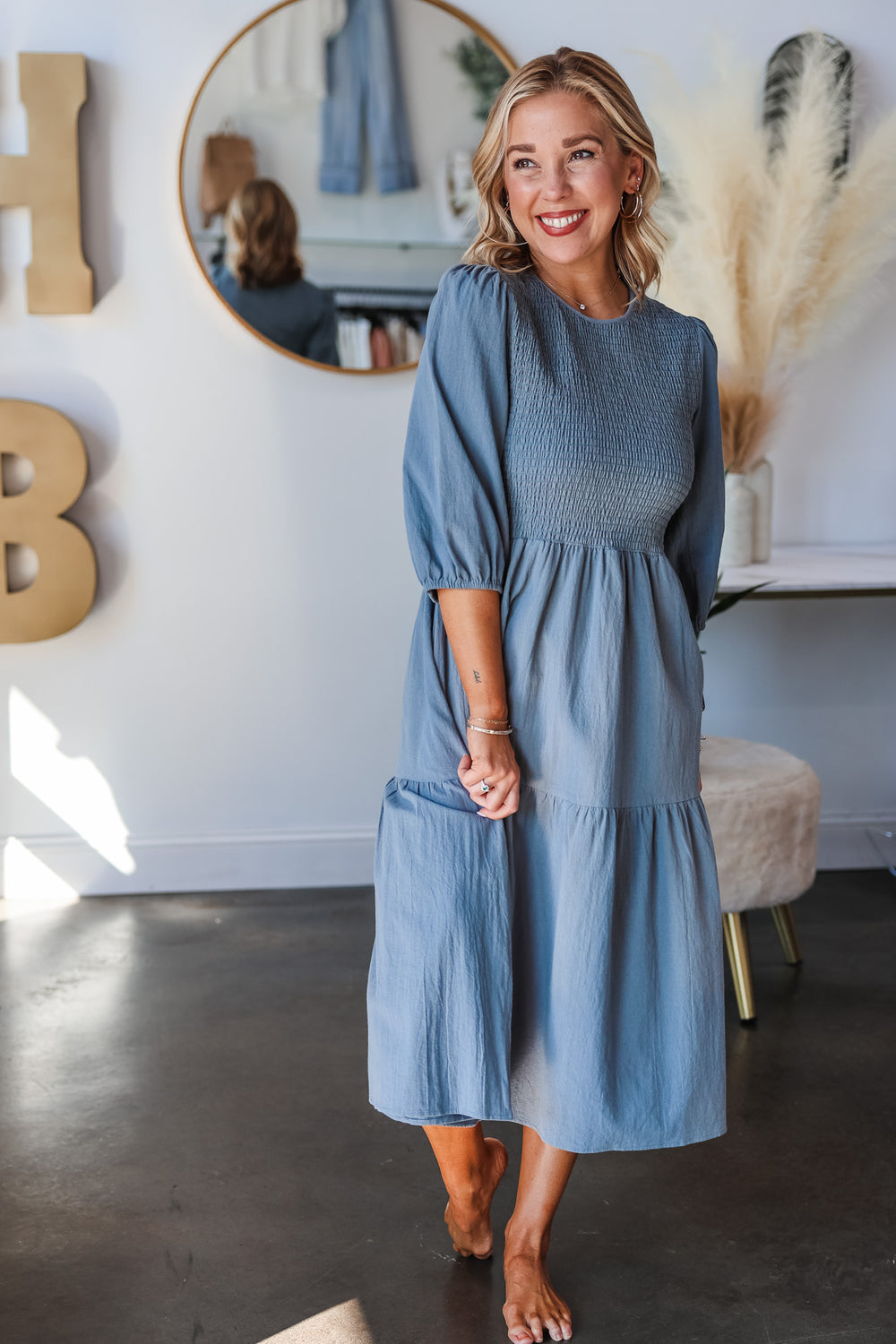 A blonde woman standing in a shop wearing a denim colored dress with 3/4 length sleeves, smocked bodice and tiered skirt.
