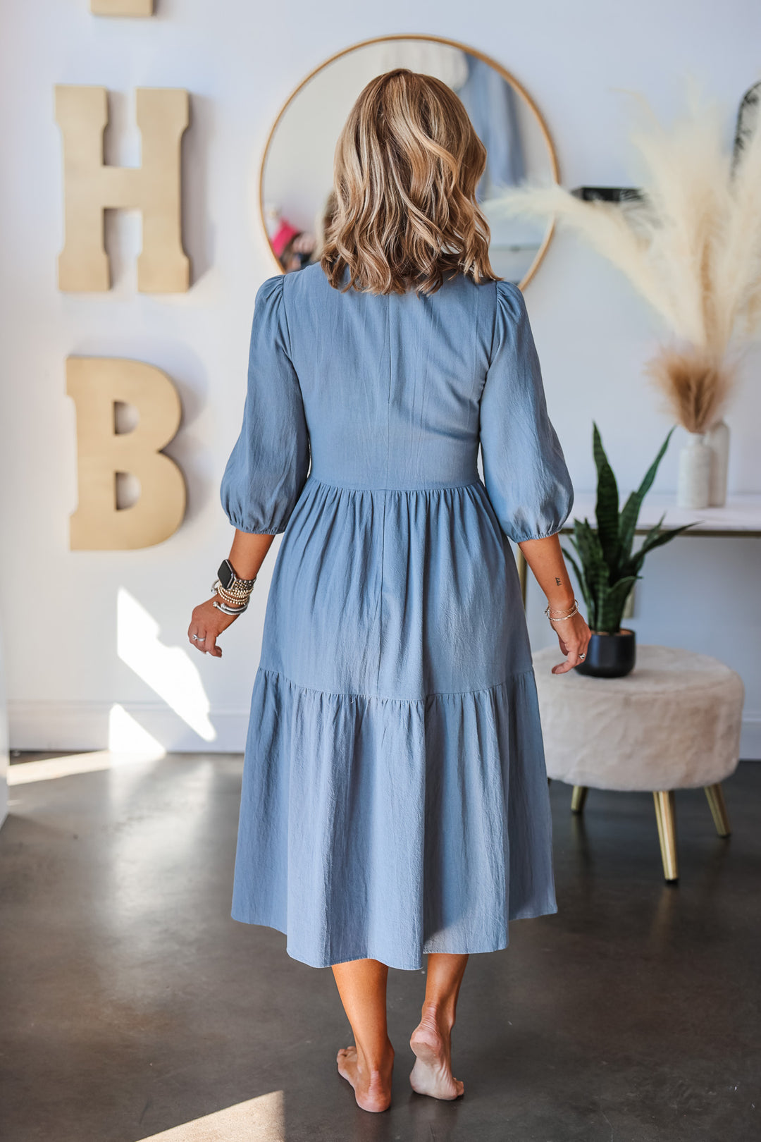 A blonde woman standing in a shop wearing a denim colored dress with 3/4 length sleeves, smocked bodice and tiered skirt. She is rear facing.