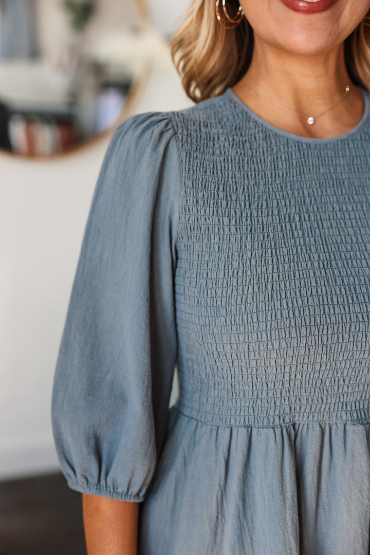 A closeup of the smocked bodice and 3/4 length sleeve on a denim colored dress.