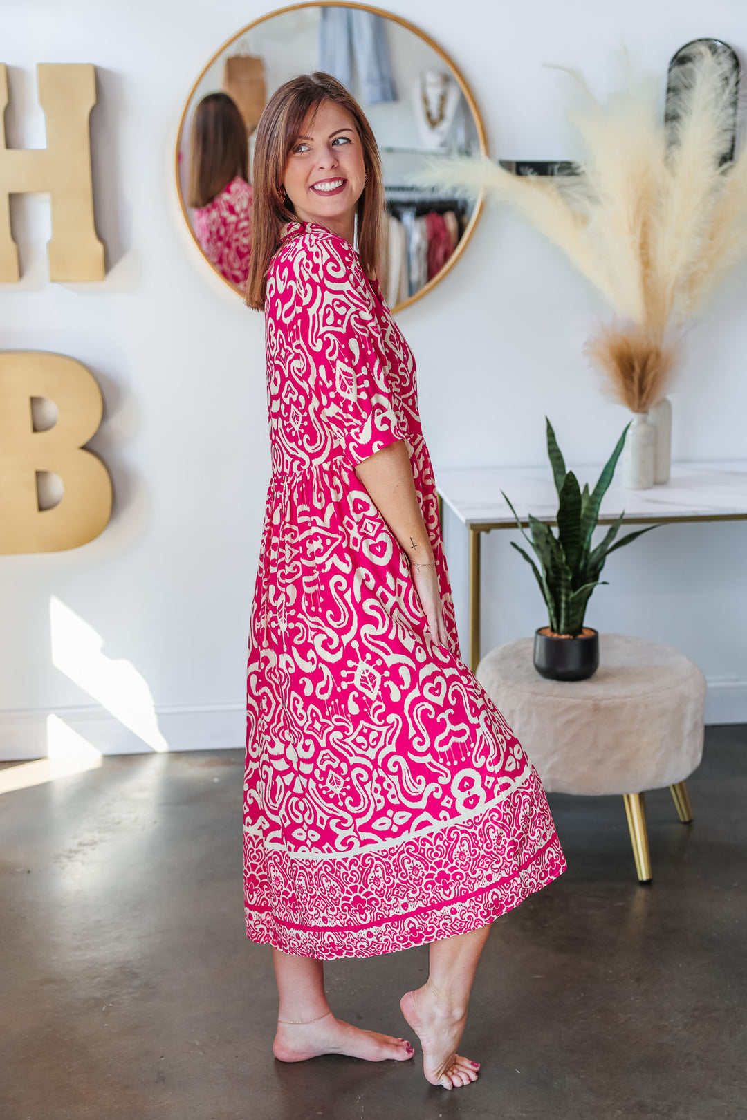 A brunette woman standing in a shop wearing a fuchsia and cream colored long dress with a button closure on top and 3/4 length sleeves.