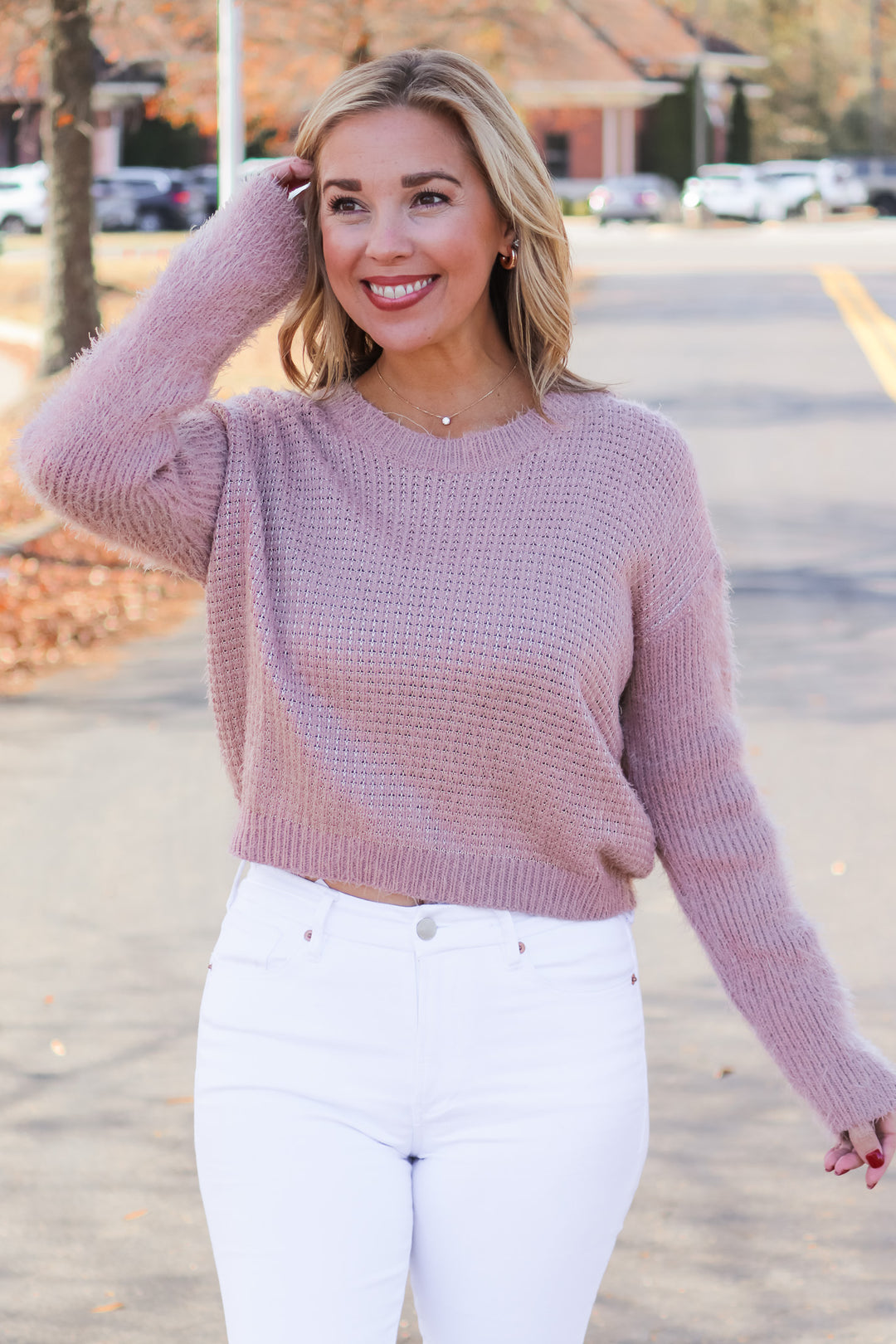 A blonde woman standing outside wearing a mauve colored long sleeve sweater and white jeans.