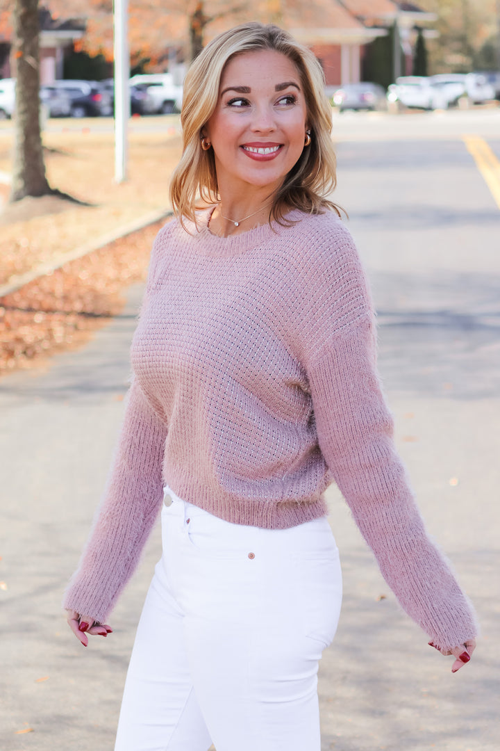 A blonde woman standing outside wearing a mauve colored long sleeve sweater and white jeans.