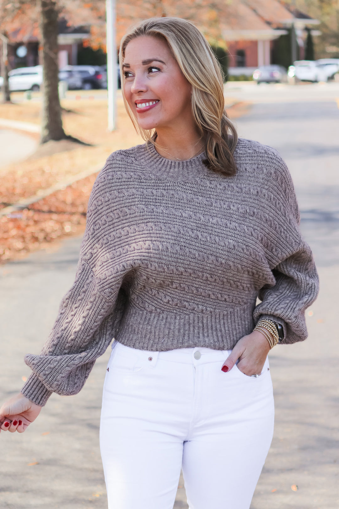 A blonde woman standing outside wearing a taupe colored cable knit sweater and white jeans.