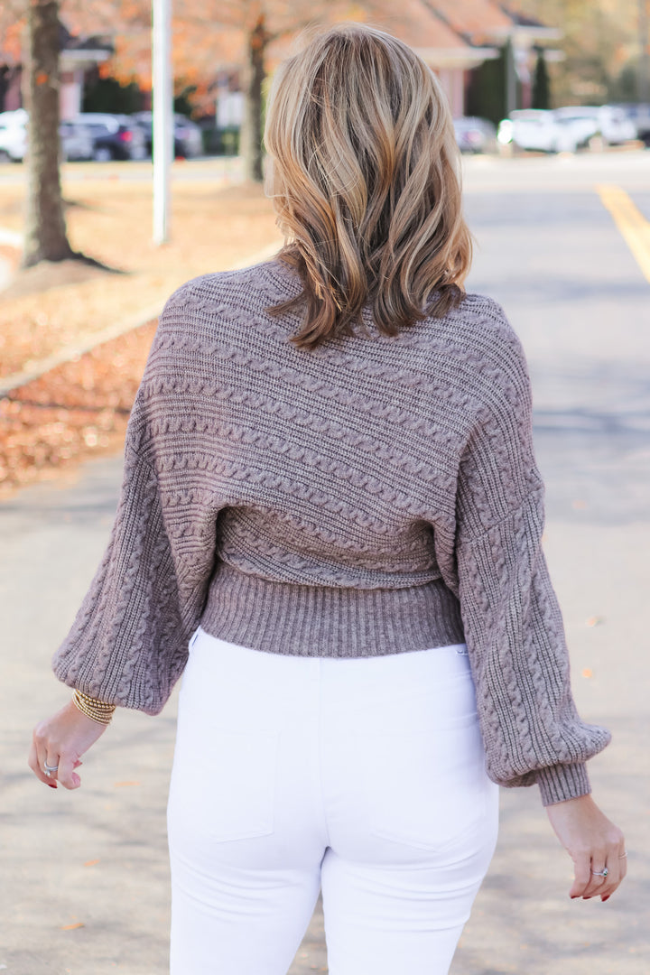 A blonde woman standing outside wearing a taupe colored cable knit sweater and white jeans. She is rear facing.