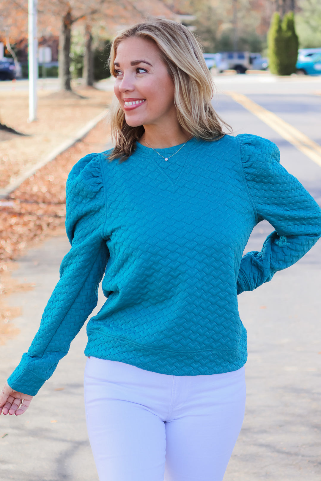A blonde woman standing outside wearing a teal quilted puff sleeve sweatshirt and white jeans.