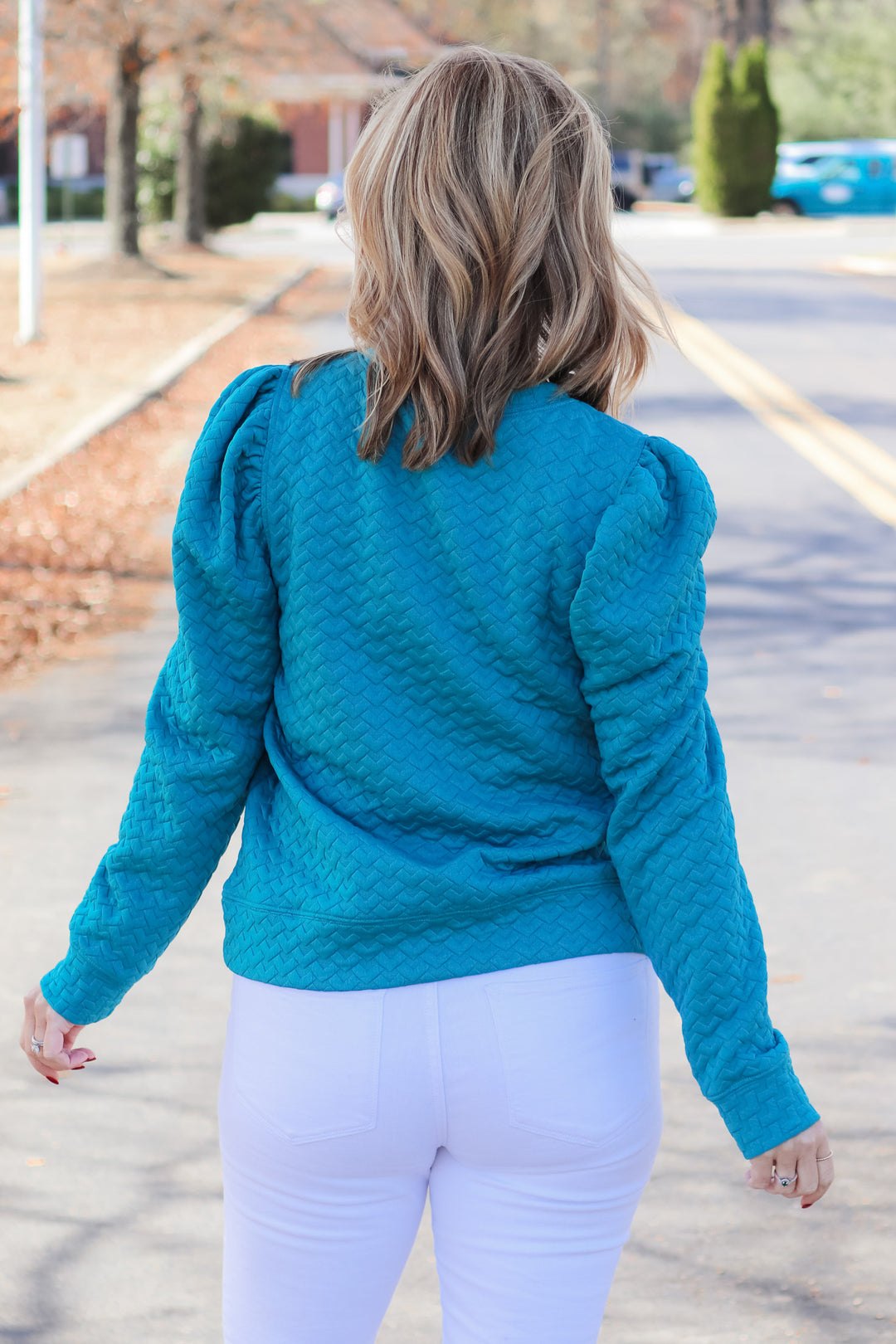 A blonde woman standing outside wearing a teal quilted puff sleeve sweatshirt and white jeans. She is rear facing.