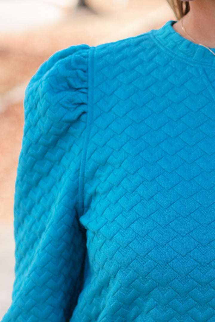 A closeup of the shoulder of a woman wearing a teal quilted puff sleeve sweatshirt.