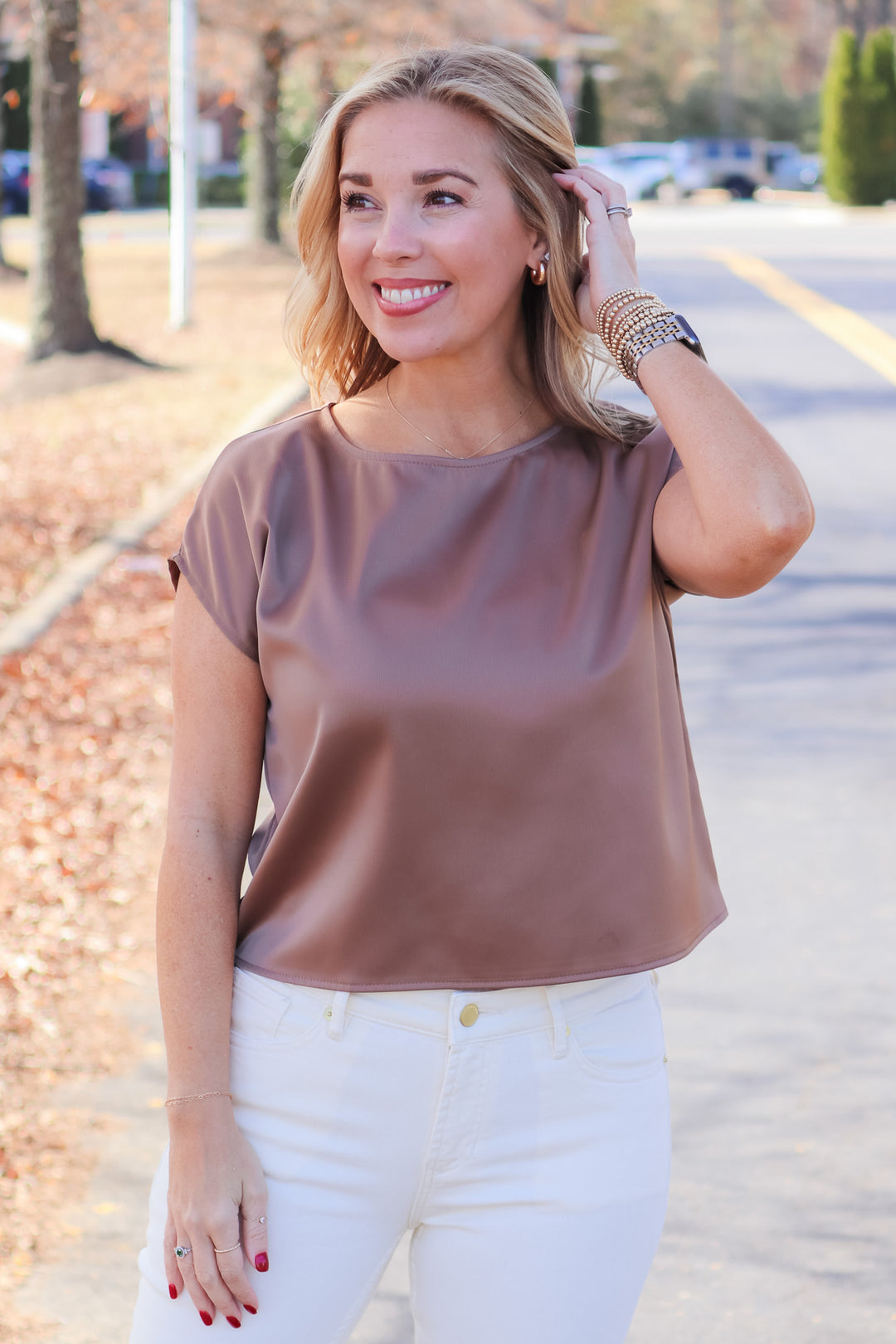 A blonde woman standing outside wearing a coffee colored satin blouse with cap sleeves and cream colored jeans.