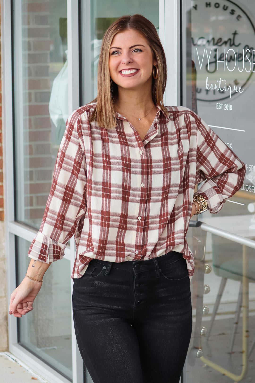 A brunette woman wearing a brown and cream colored plaid button down top. She is standing in front of a shop.