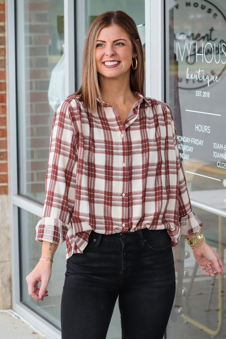 A brunette woman wearing a brown and cream colored plaid button down top. She is standing in front of a shop.