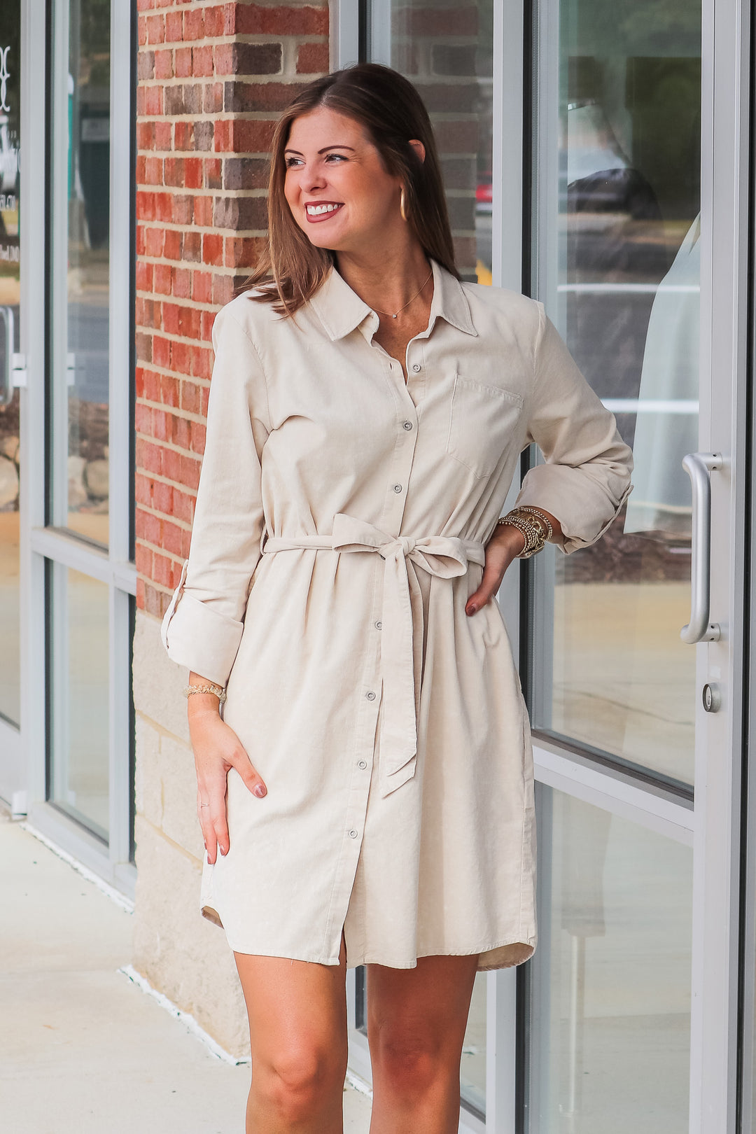 A brunette woman wearing a tan corduroy shirt dress with a tie belt. She is standing in front of a shop.