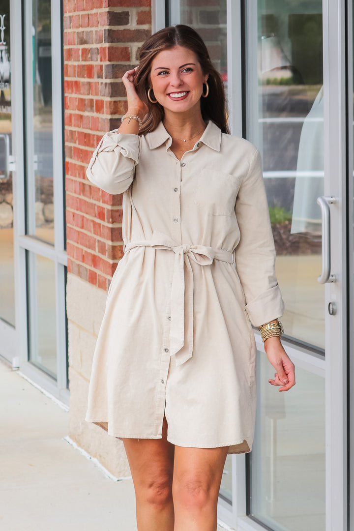 A brunette woman wearing a tan corduroy shirt dress with a tie belt. She is standing in front of a shop.