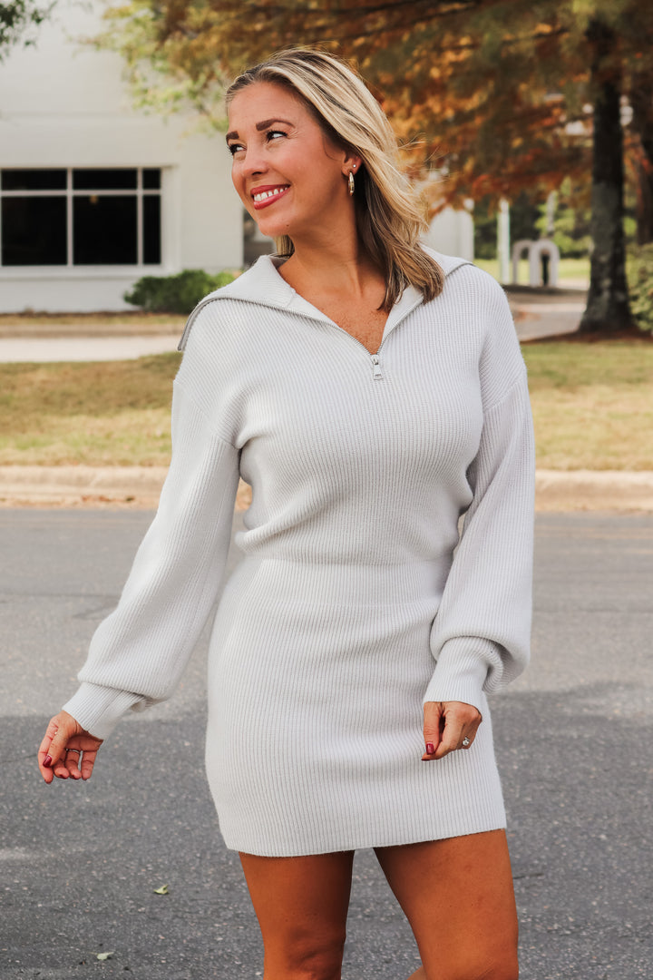 A blonde woman wearing a very light gray sweater dress. It is fitted with an oversized collar that zips. She is standing outside.  