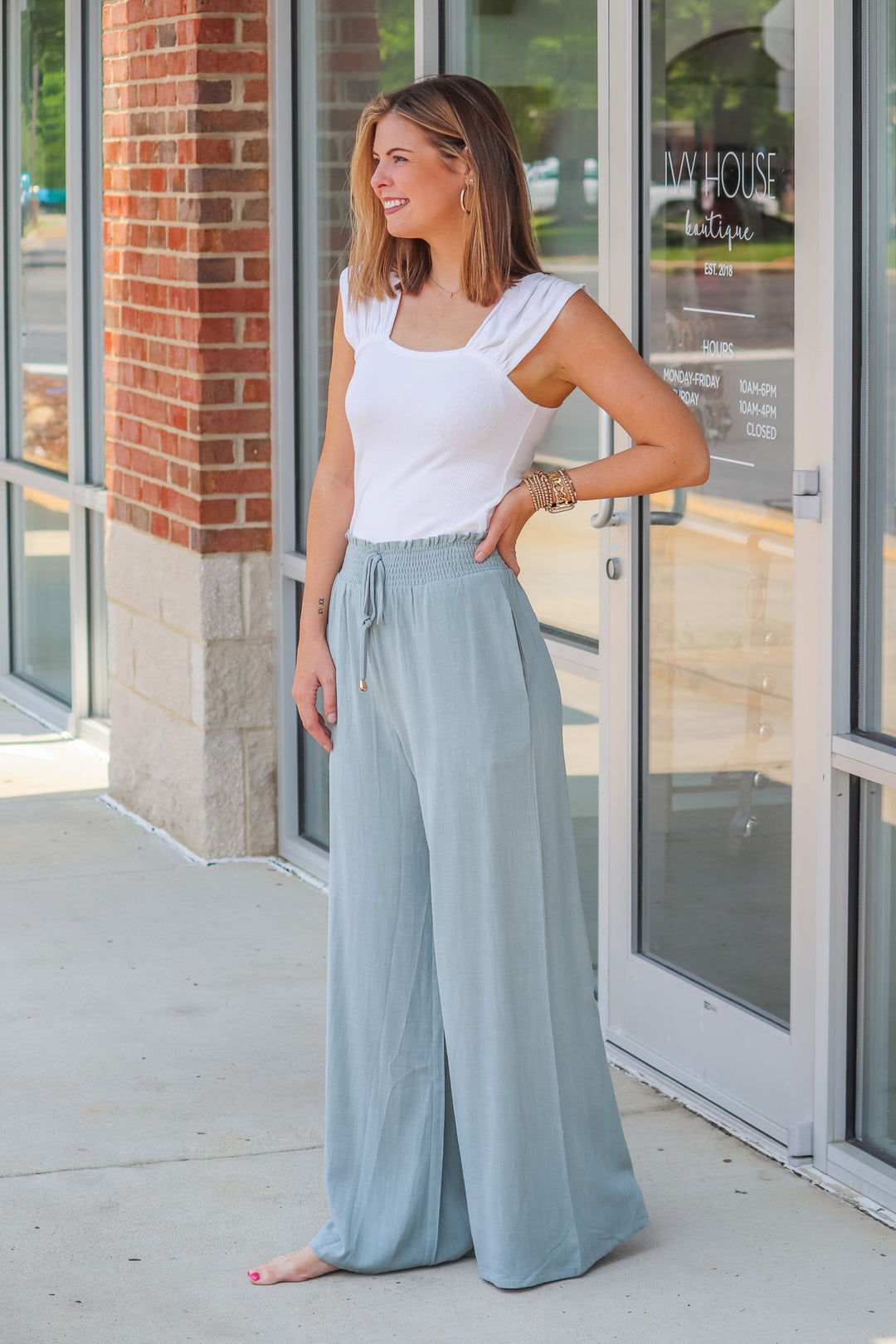 A woman wearing sage colored wide leg linen pants with a drawstring and elastic waistband. She is standing in front of a shop.  