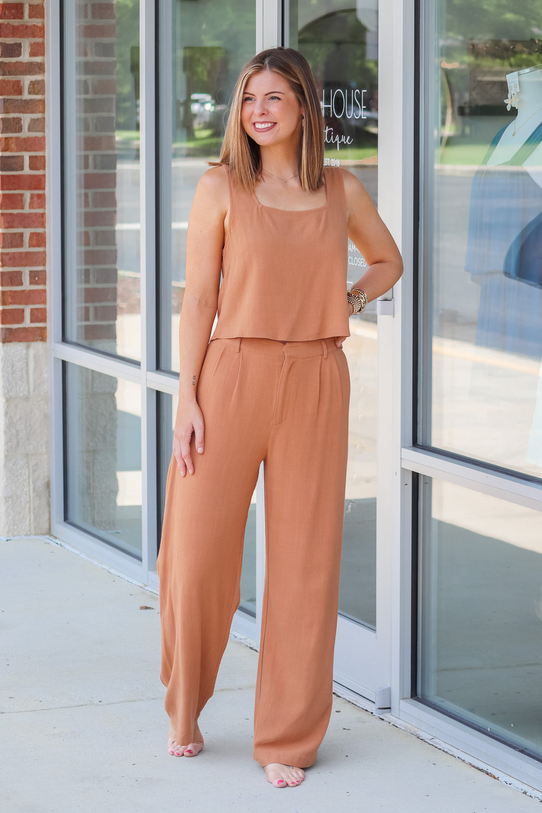 A woman wearing a tan top and tan linen pants with belt loops and pockets. She is standing in front of s shop.
