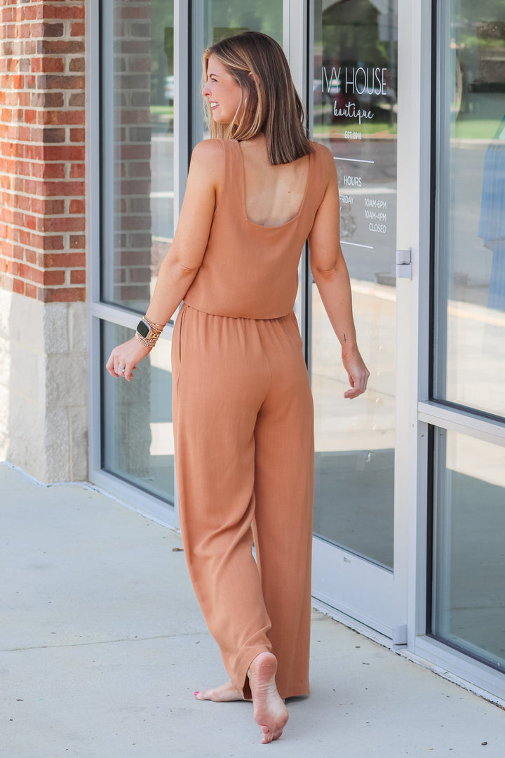 A brunette woman wearing a tan, square neckline, waist length top and matching pants. She is standing in front of a shop.  She is rear facing.