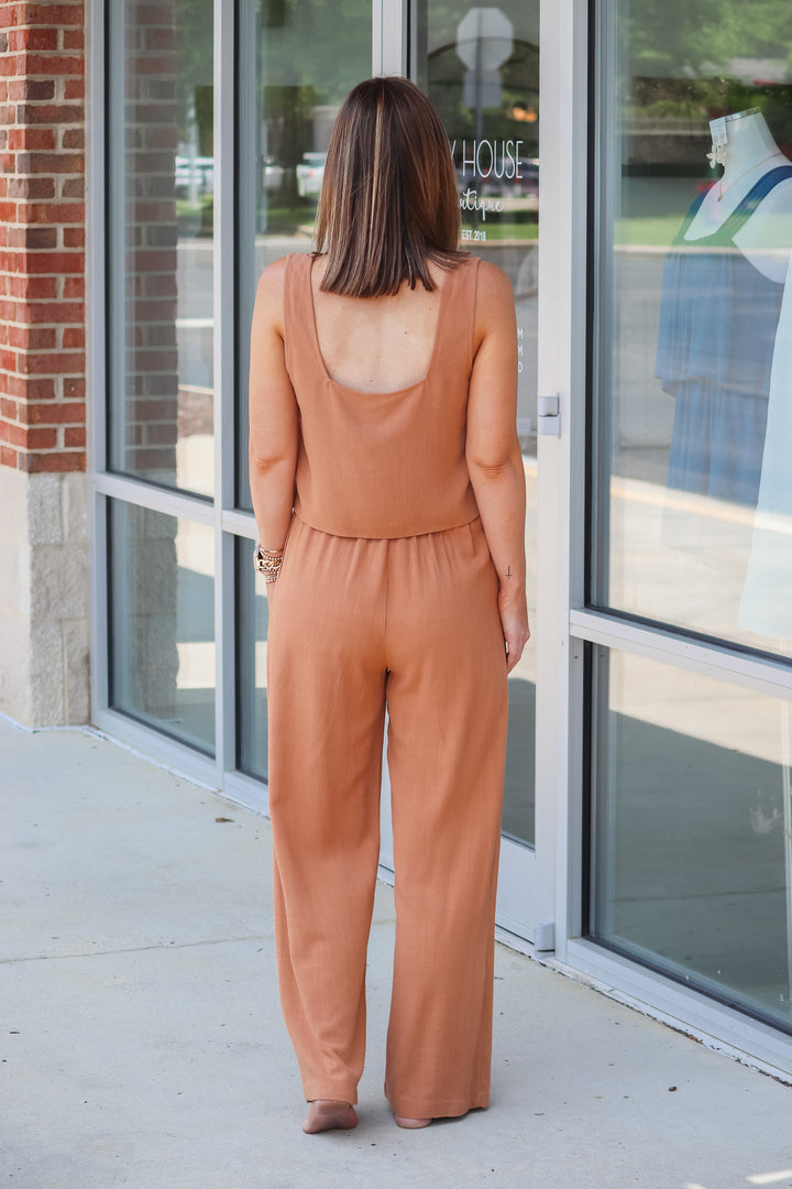 A woman wearing a tan top and tan linen pants with belt loops and pockets. She is standing in front of s shop. She is rear facing.