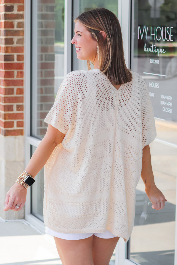 A brunette woman wearing a cream colored short sleeve crochet sweater and white tank. She is standing in front of a shop. She is rear facing.