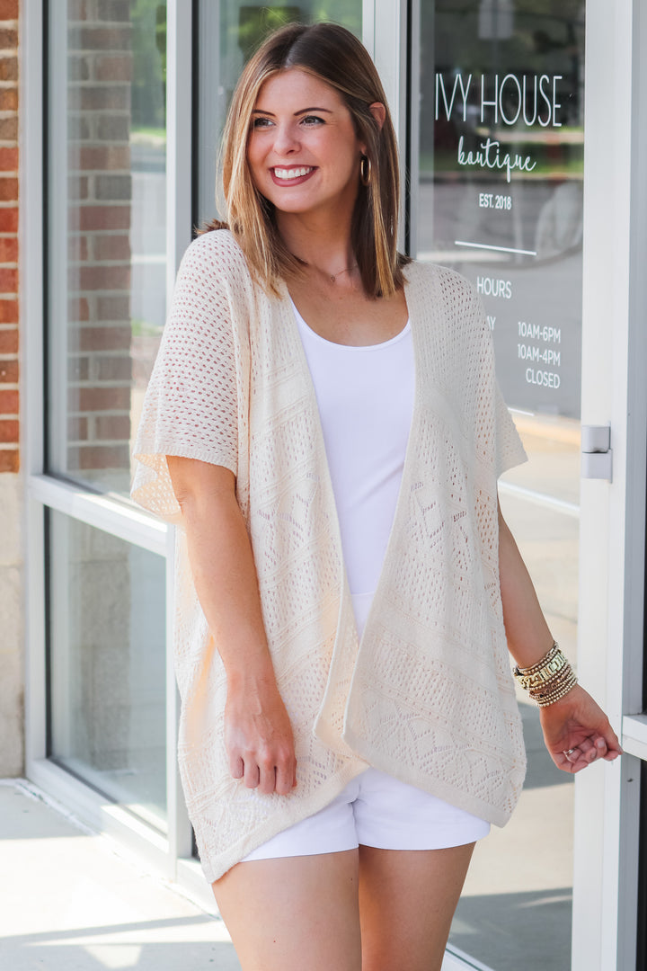 A brunette woman wearing a cream colored short sleeve crochet sweater and white tank. She is standing in front of a shop.