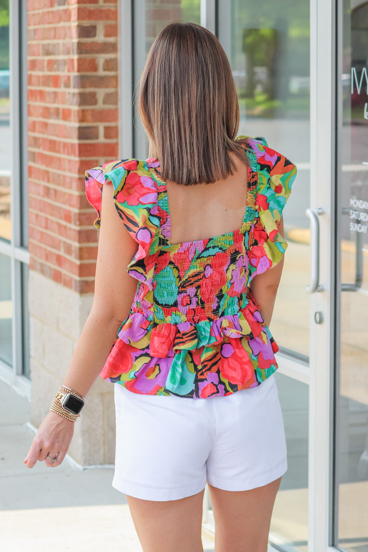 A brunette woman wearing a colorful smocked top with a ruffle peplum hem and ruffle shoulders. The colors are red, black, teal, yellow, purple and green. She is standing in front of a shop. She is rear facing.