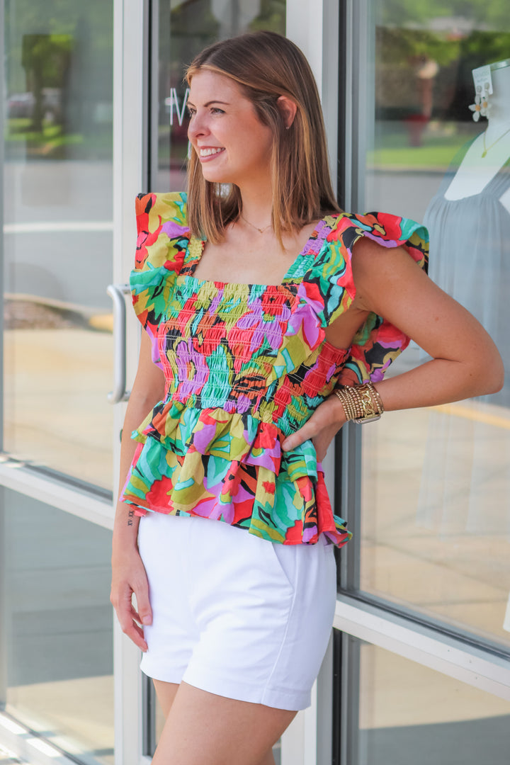 A brunette woman wearing a colorful smocked top with a ruffle peplum hem and ruffle shoulders. The colors are red, black, teal, yellow, purple and green. She is standing in front of a shop.