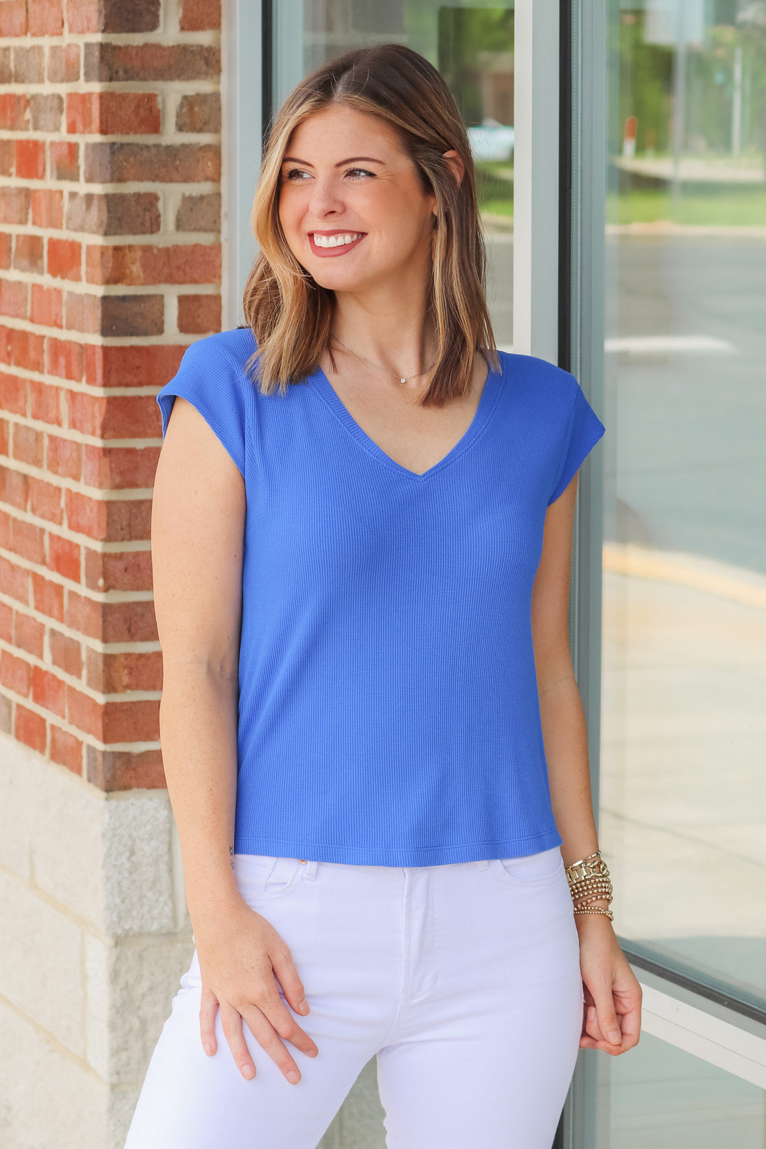 A brunette woman wearing a blue shirt with cap sleeves and a v neck. She is standing in front of a shop.  