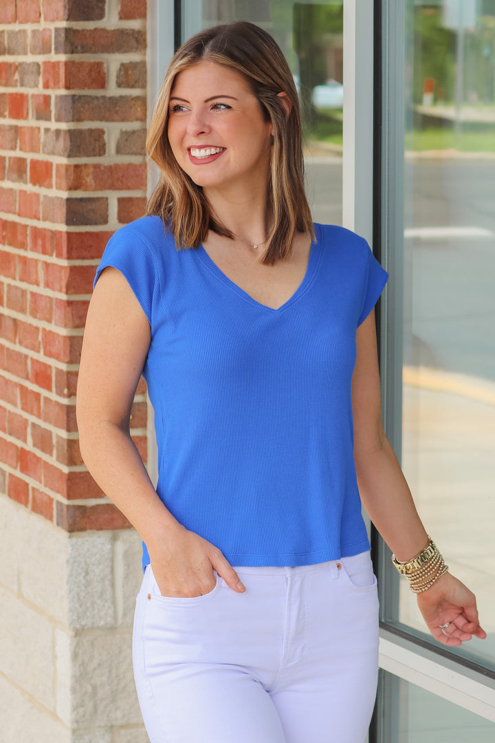 A brunette woman wearing a blue shirt with cap sleeves and a v neck. She is standing in front of a shop.