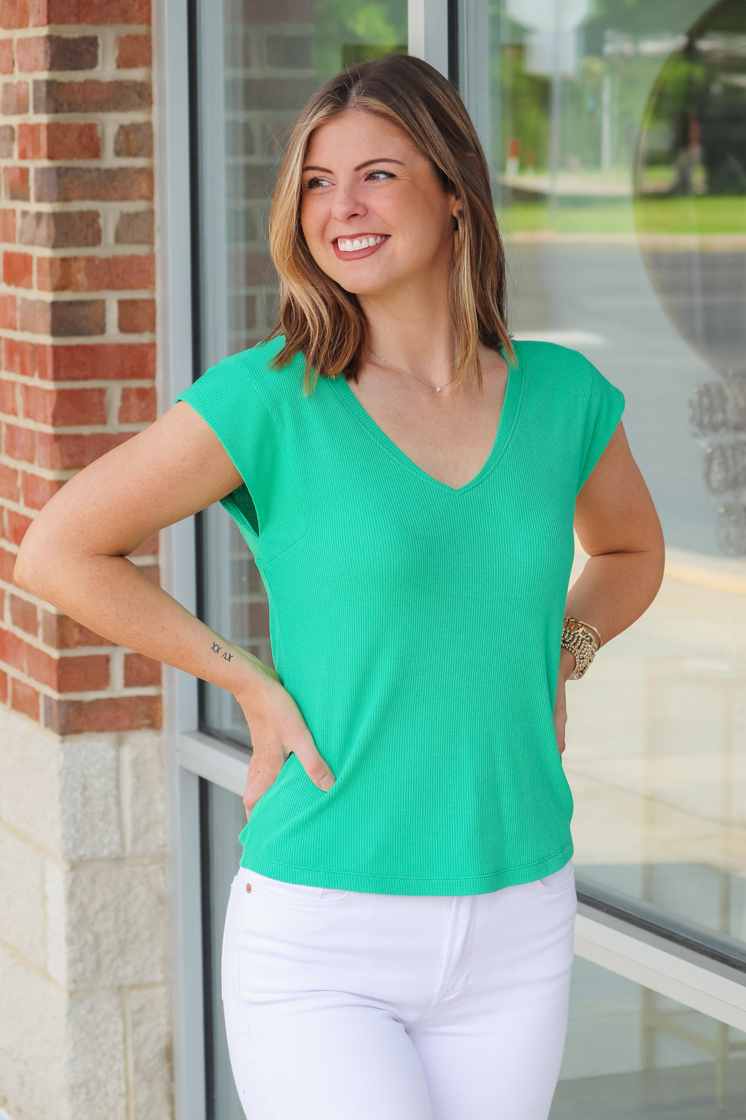 A brunette woman wearing a green shirt with cap sleeves and a v neck. She is standing in front of a shop.