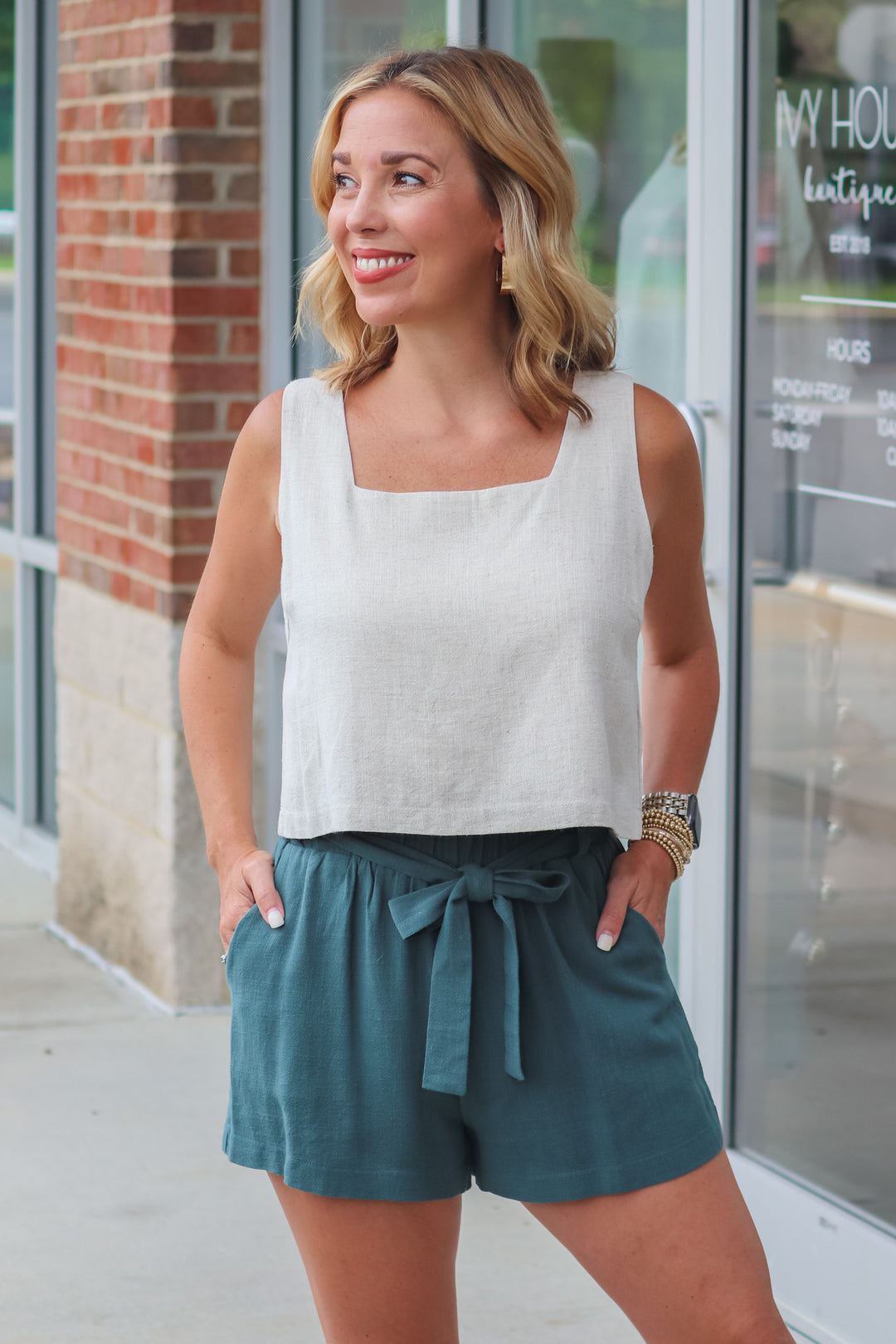 A woman wearing green linen shorts with an elastic waist and a tie belt. She is standing in front of a shop.