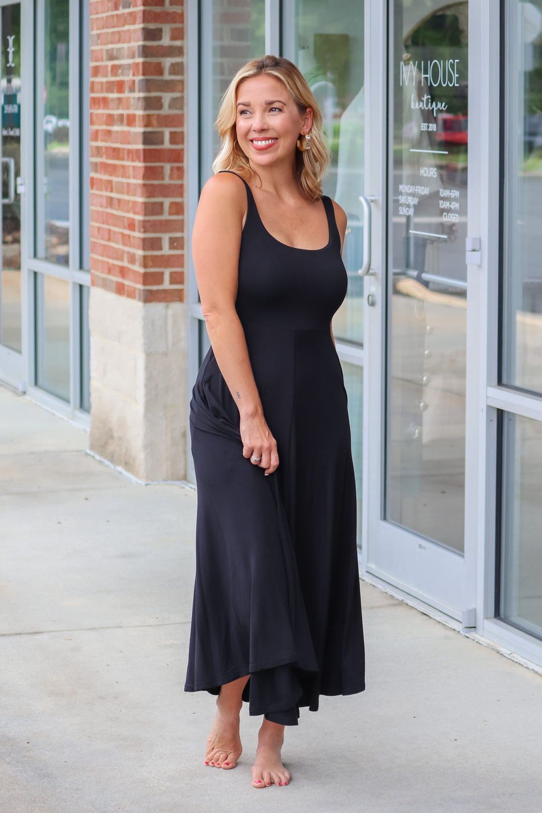 A blonde woman wearing a black maxi dress with a square neckline and pockets. She is standing in front of a shop.