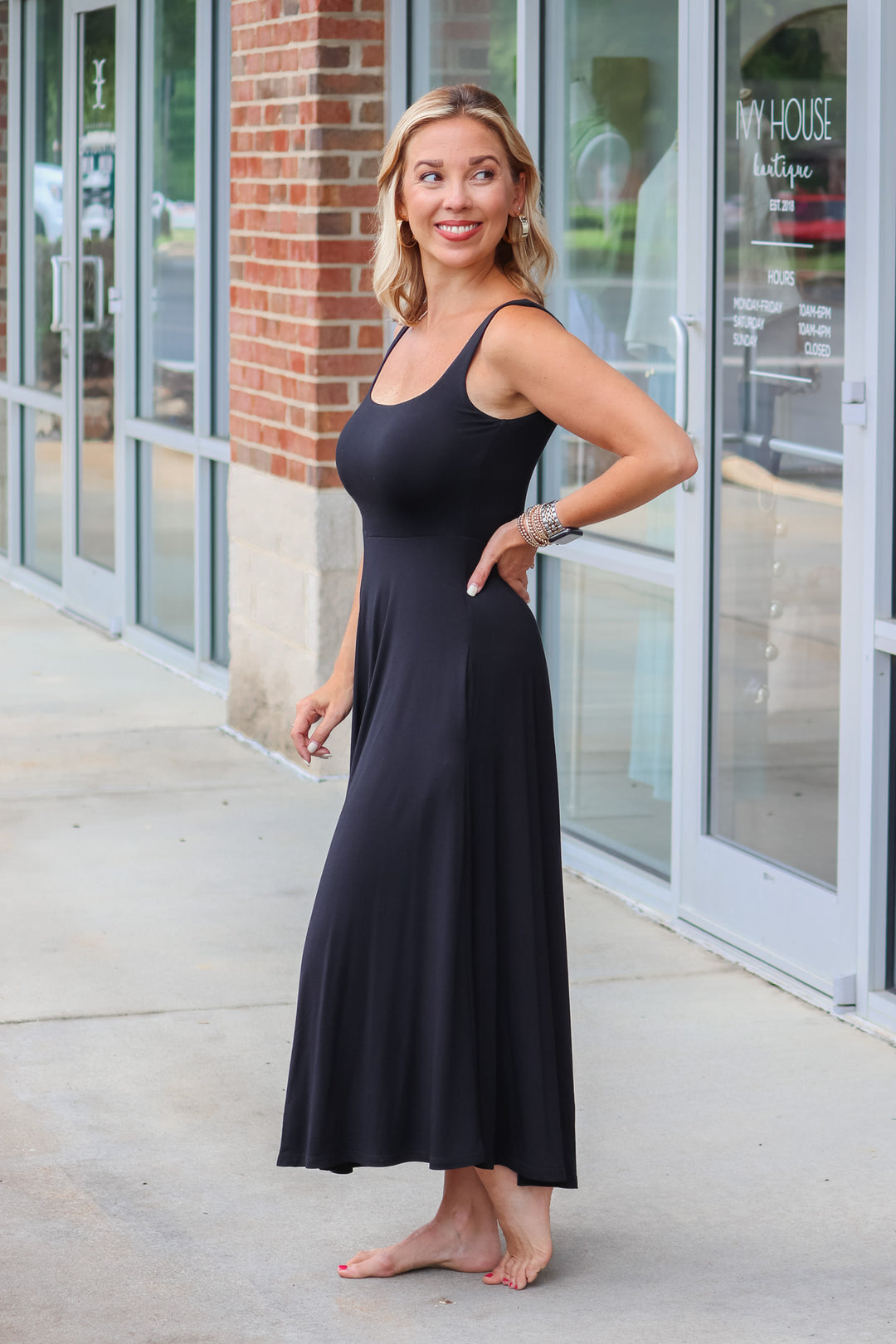 A blonde woman wearing a black maxi dress with a square neckline and pockets. She is standing in front of a shop.  