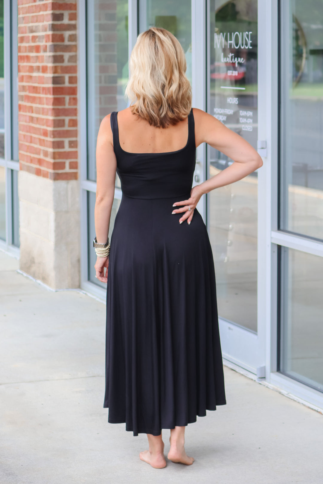 A blonde woman wearing a black maxi dress with a square neckline and pockets. She is standing in front of a shop.  She is rear facing.