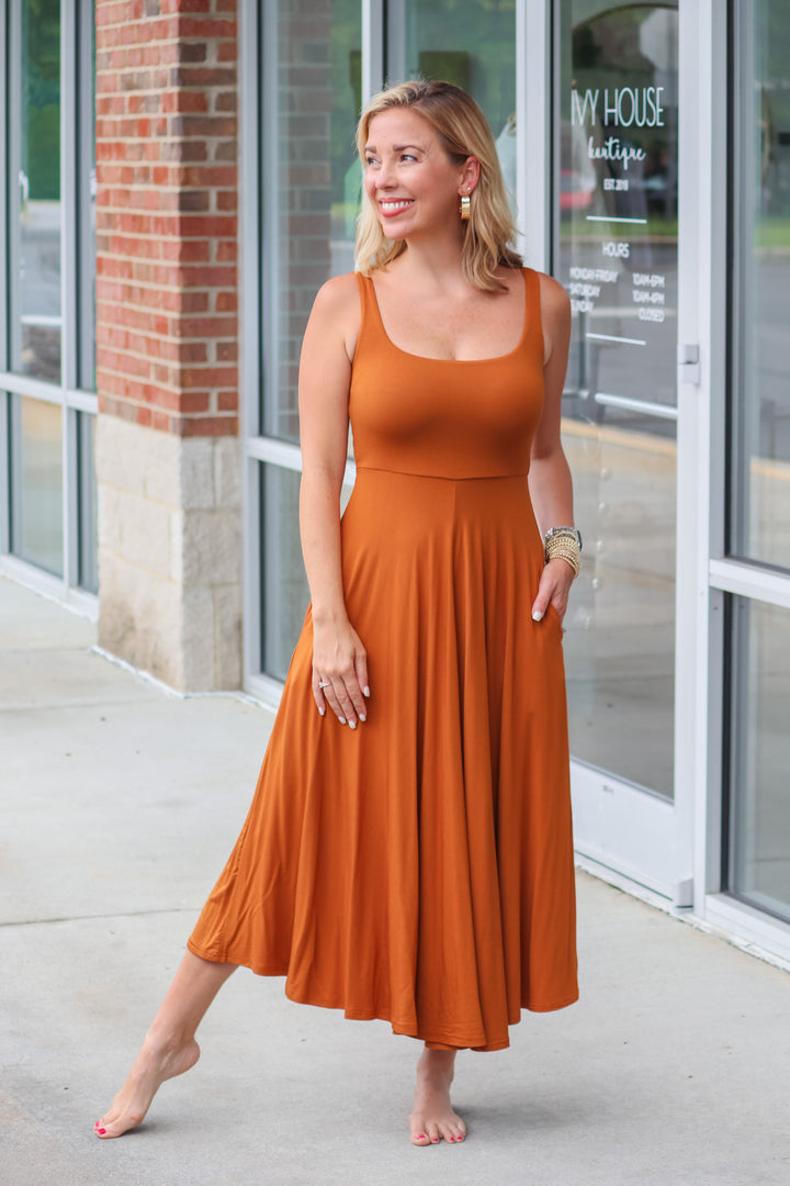 A blonde woman wearing an almond colored maxi dress with a square neckline and pockets. She is standing in front of a store.