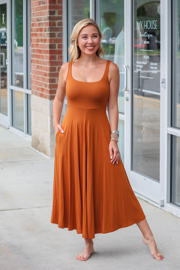 A blonde woman wearing an almond colored maxi dress with a square neckline and pockets. She is standing in front of a shop.