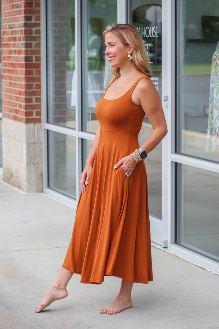 A blonde woman wearing an almond colored maxi dress with a square neckline and pockets. She is standing in front of a shop.
