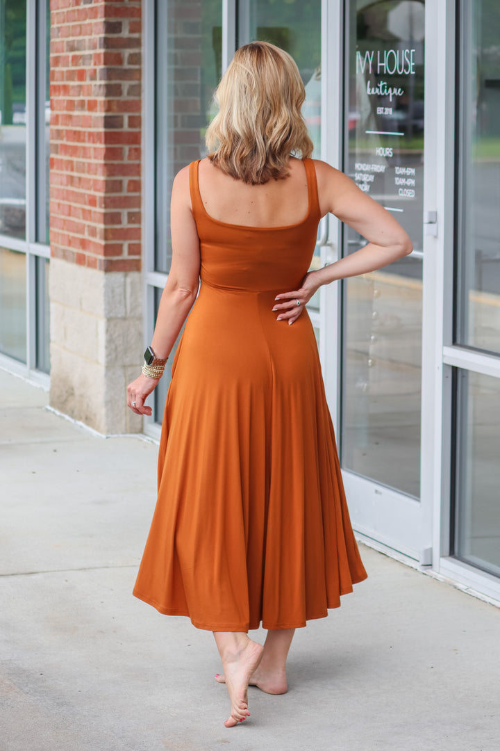 A blonde woman wearing an almond colored maxi dress with a square neckline and pockets. She is standing in front of a shop. She is rear facing.
