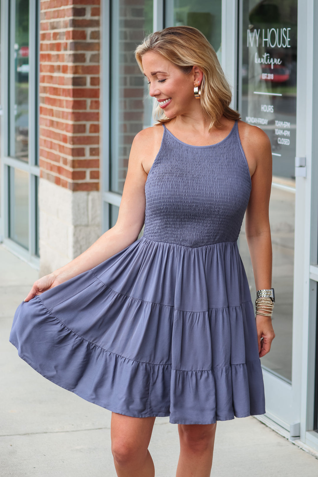 A blonde woman wearing a navy blue dress with a smocked top and tiered skirt. She is standing in front of a shop.