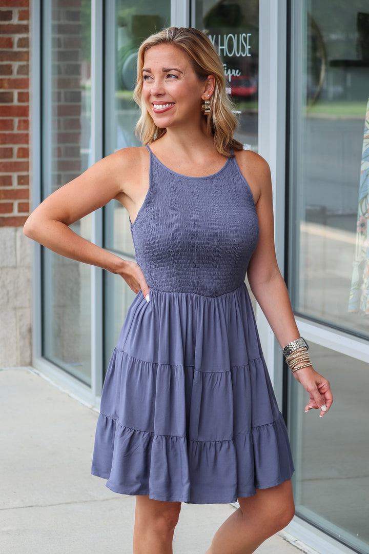 A blonde woman wearing a navy blue dress with a smocked top and tiered skirt. She is standing in front of a shop.