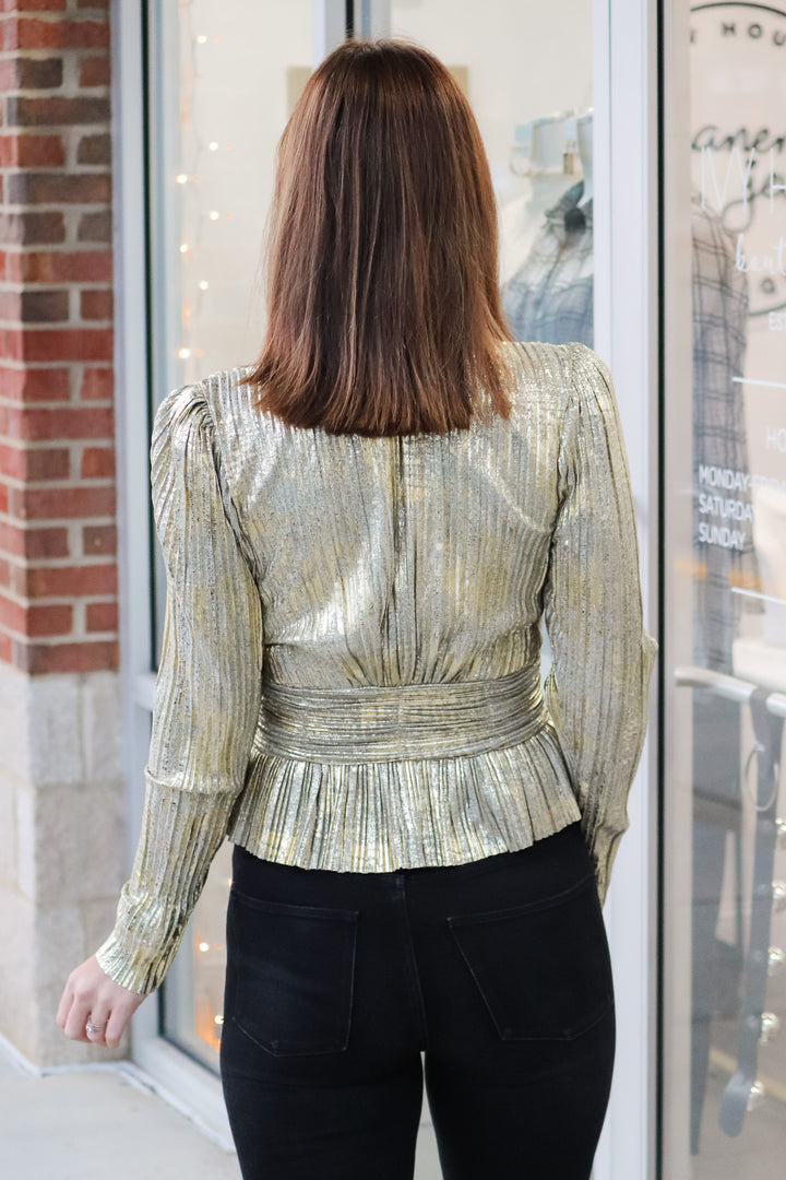 A brunette woman standing in front of a shop wearing a metallic gold pleated top with long sleeves, peplum hem and a v neck with black jeans. She is rear facing.