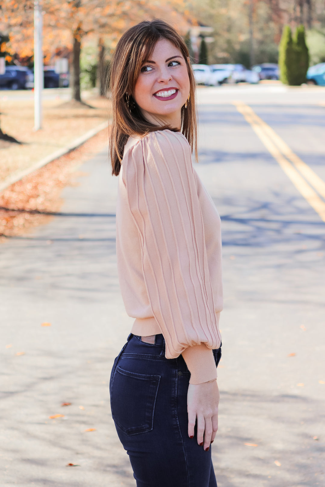 A brunette woman standing outside wearing an oat colored detailed long sleeve sweater with blue jeans.