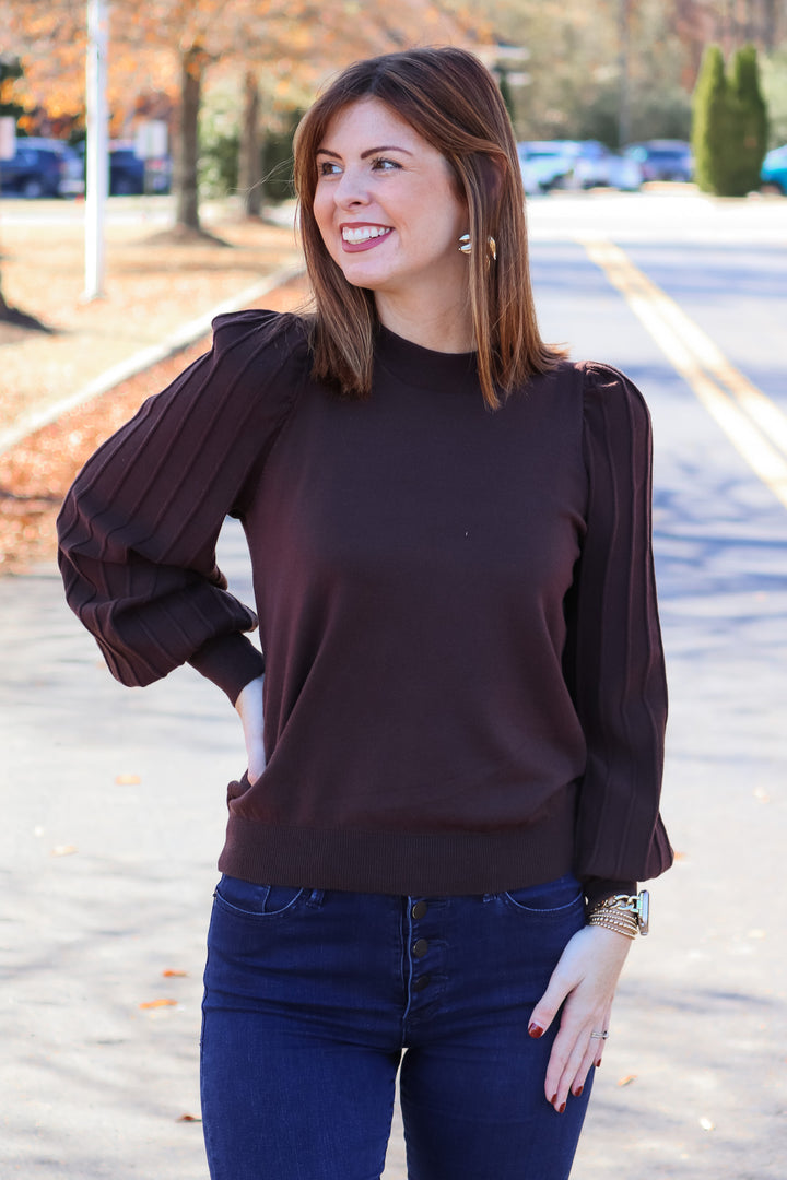 A brunette woman standing outside wearing a brown sweater with long detailed sleeves and blue jeans.