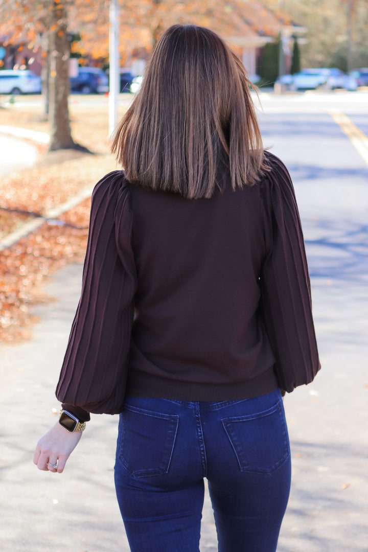 A brunette woman standing outside wearing a brown sweater with long detailed sleeves and blue jeans. She is rear facing.
