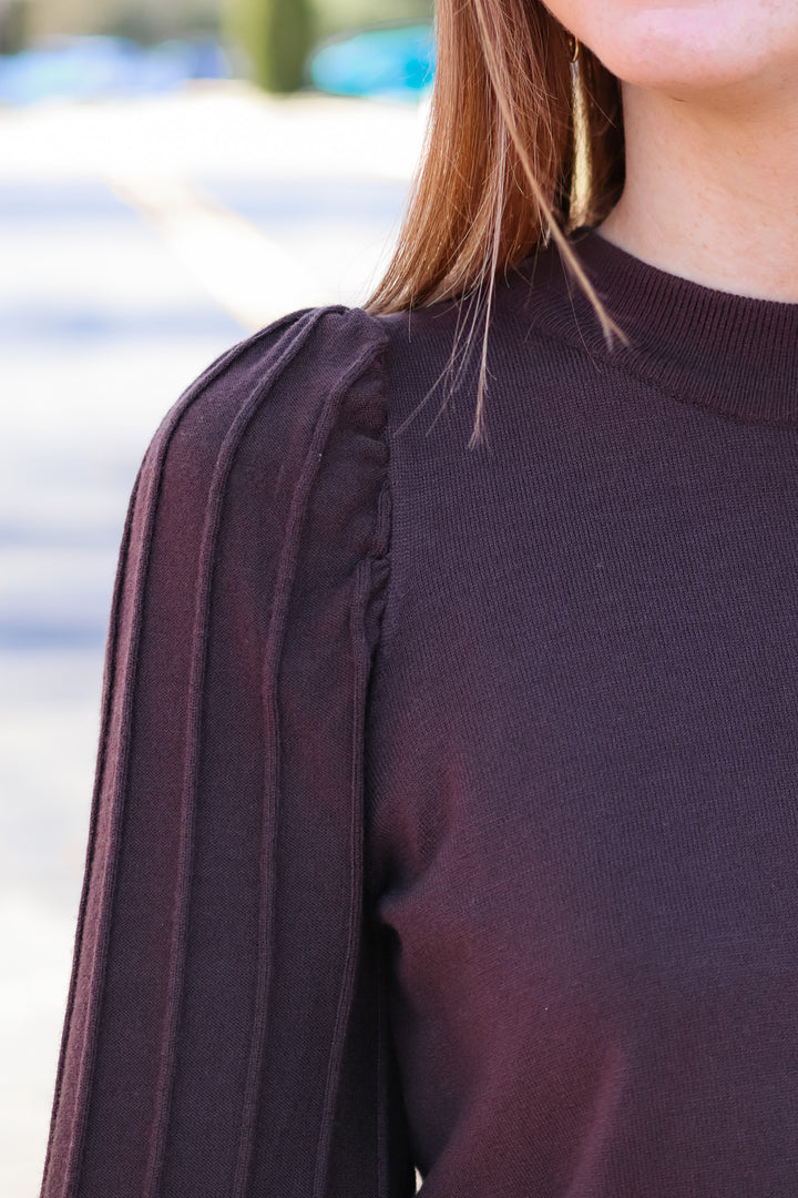 A closeup of the shoulder of a woman wearing a brown detailed sleeve sweater.