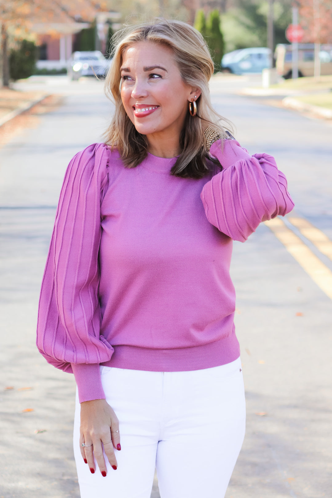 A blonde woman standing outside wearing a pink detailed long sleeve sweater and white jeans.