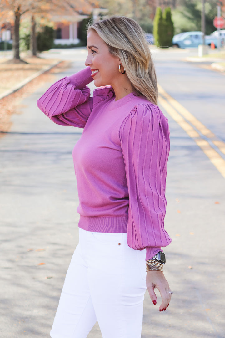 A blonde woman standing outside wearing a pink detailed long sleeve sweater and white jeans.