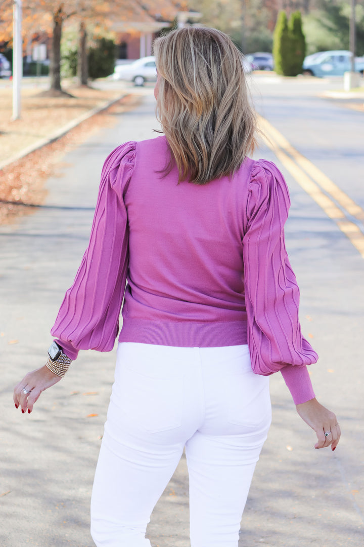A blonde woman standing outside wearing a pink detailed long sleeve sweater and white jeans. She is rear facing.
