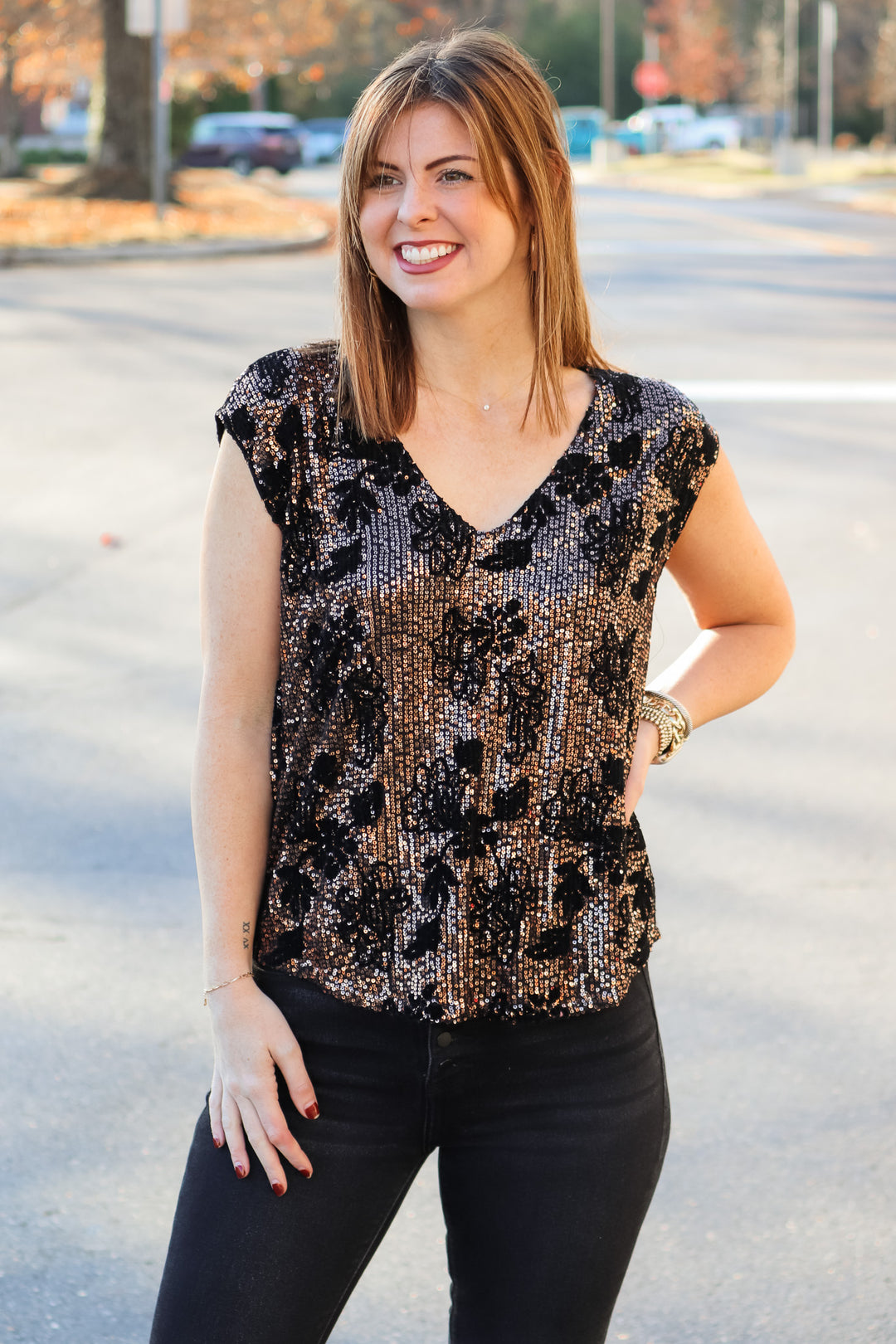 A brunette woman standing outside wearing a cap sleeve rose gold and black sequin top with a v neck and black jeans.